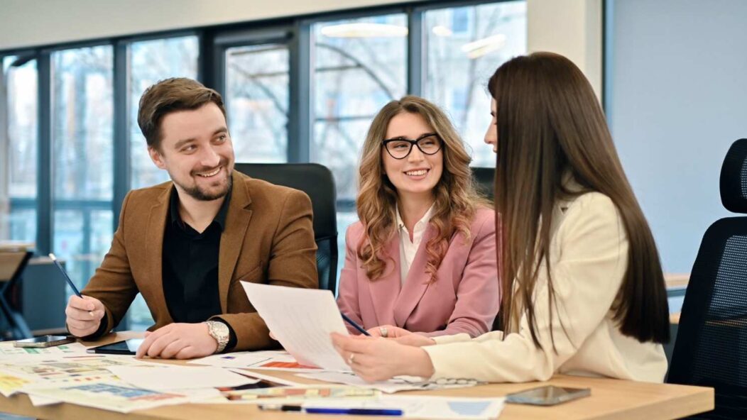 A diverse team of three people sitting around a table, working on travel documents in a modern office environment, with a globe and a travel agency flyer on the table.