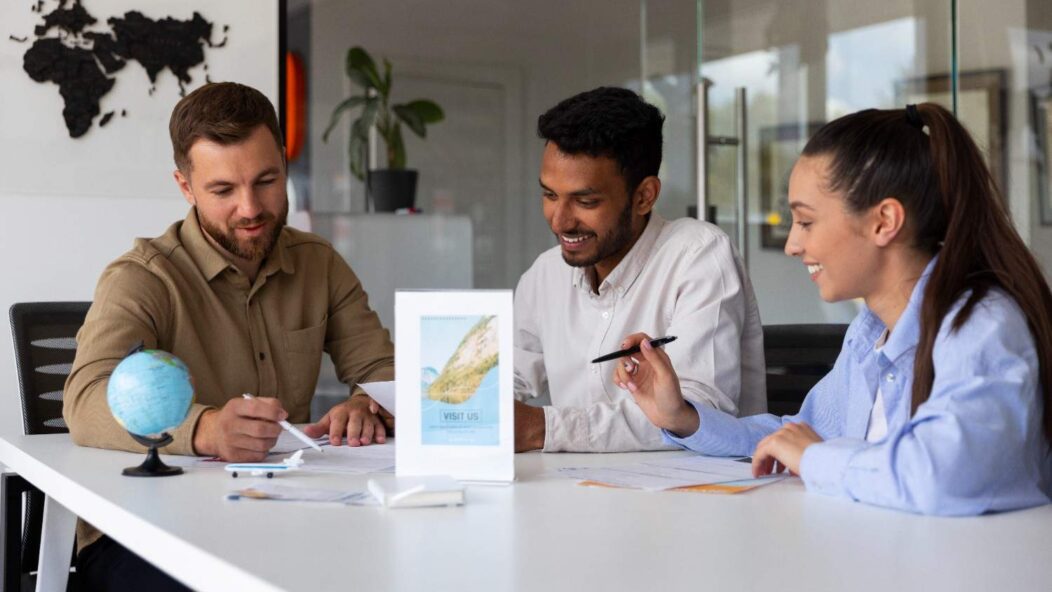 A diverse team of three people sitting around a table, working on travel documents in a modern office environment, with a globe and a travel agency flyer on the table.