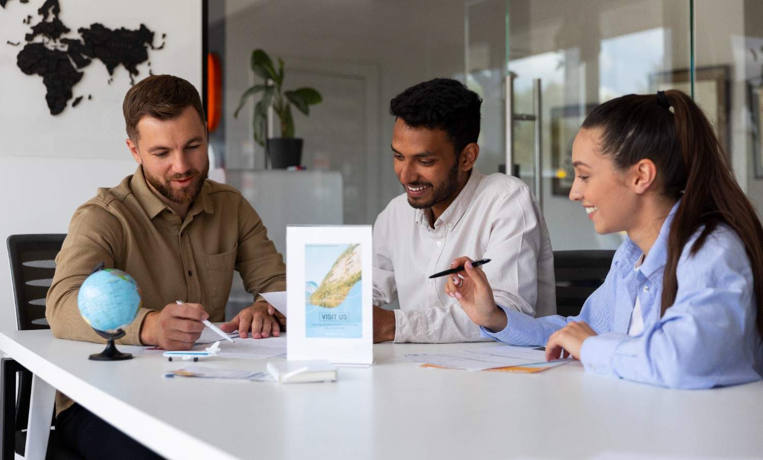 A diverse team of three people sitting around a table, working on travel documents in a modern office environment, with a globe and a travel agency flyer on the table.