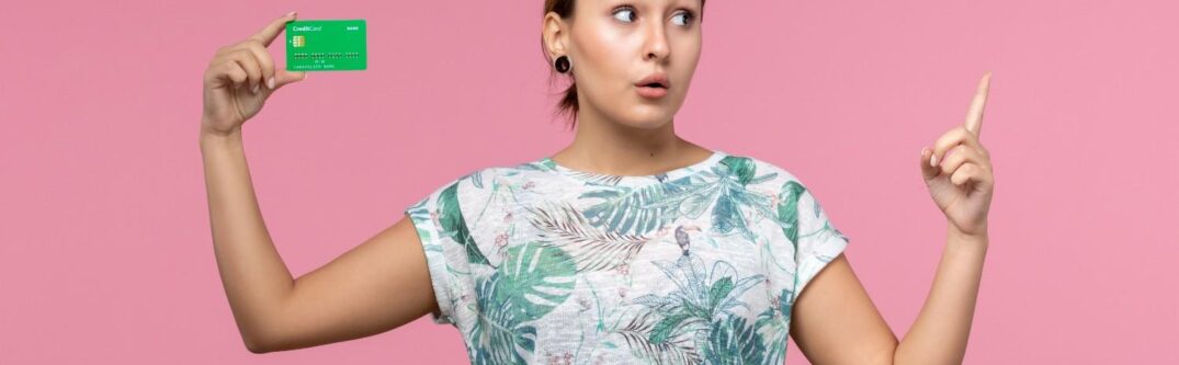 Young woman holding a green card with a suitcase, appearing thoughtful against a pink background – concept of green card renewal and travel.