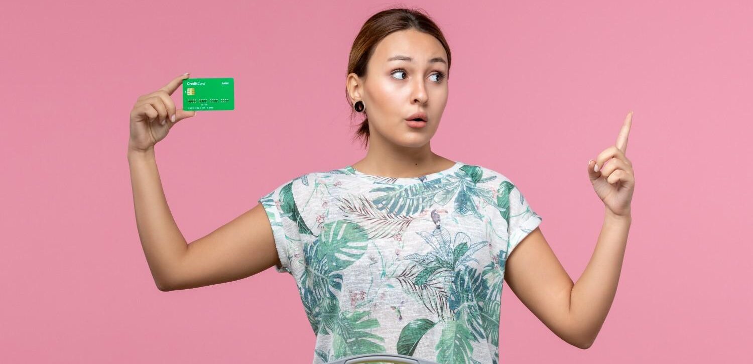 Young woman holding a green card with a suitcase, appearing thoughtful against a pink background – concept of green card renewal and travel.