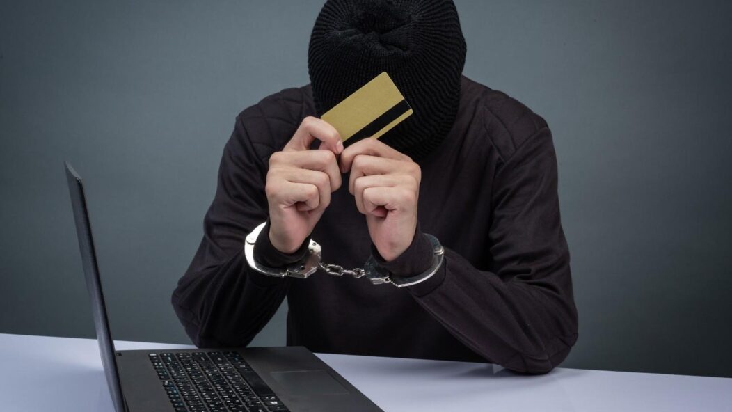 H-1B visa fraud concept with a man in handcuffs holding a credit card in front of a laptop, symbolizing illegal practices and consequences.