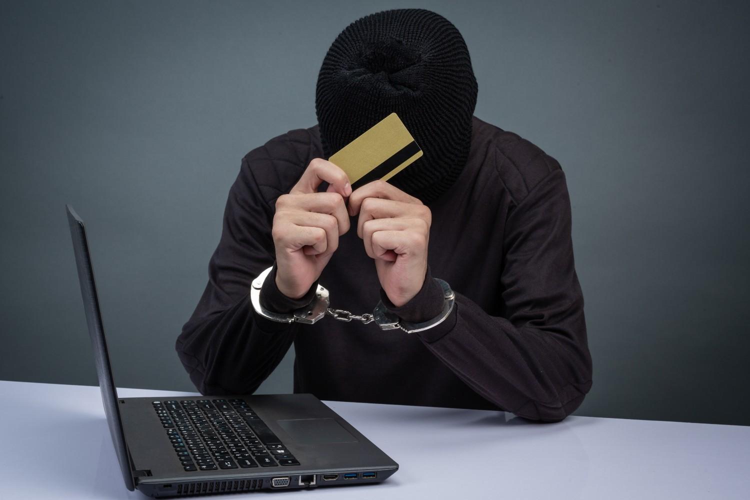 H-1B visa fraud concept with a man in handcuffs holding a credit card in front of a laptop, symbolizing illegal practices and consequences.