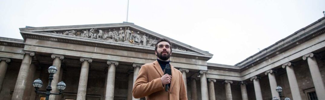 Man in a brown coat holding a microphone in front of a historical building with columns, symbolizing authority and public speaking.