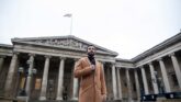 Man in a brown coat holding a microphone in front of a historical building with columns, symbolizing authority and public speaking.