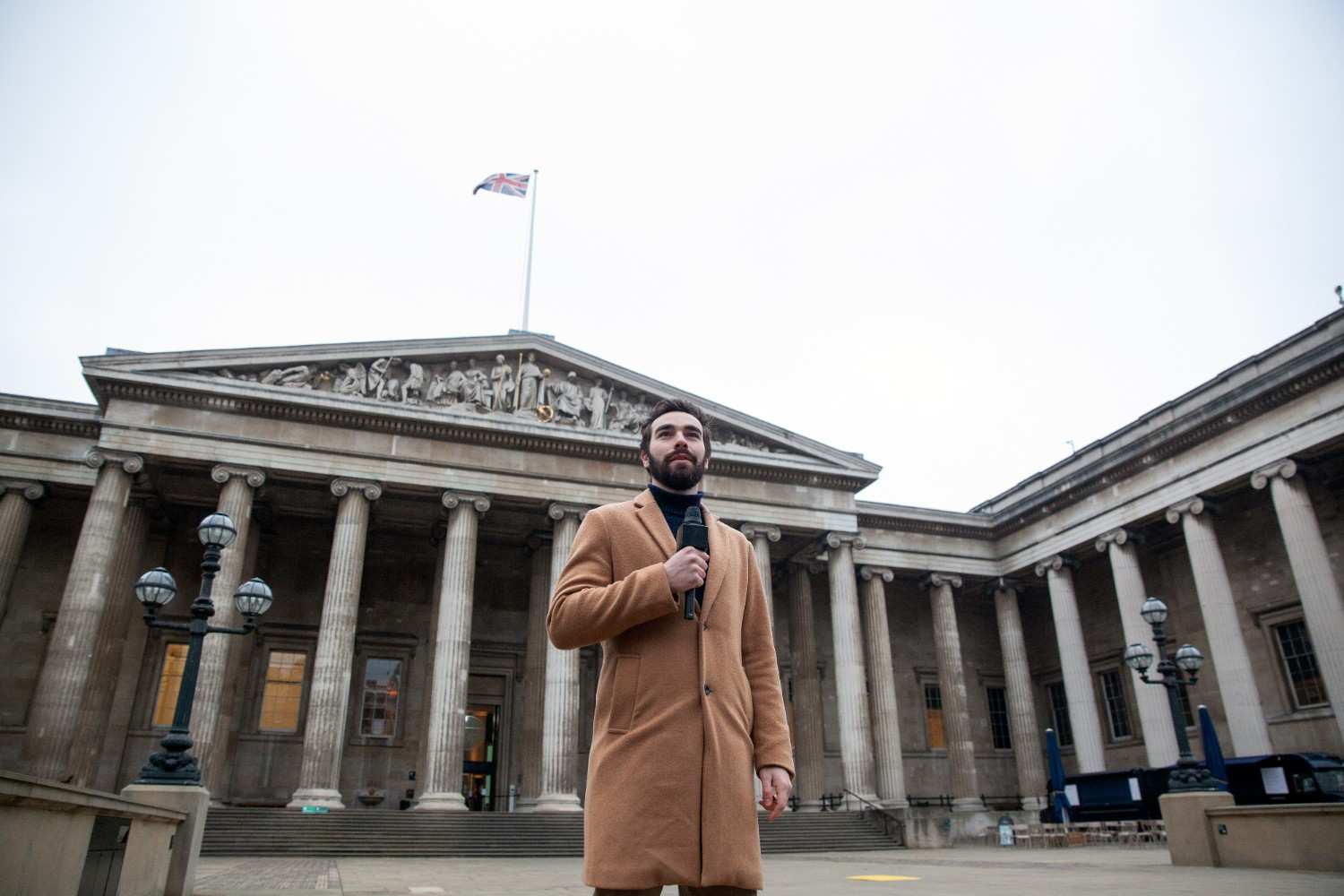 Man in a brown coat holding a microphone in front of a historical building with columns, symbolizing authority and public speaking.