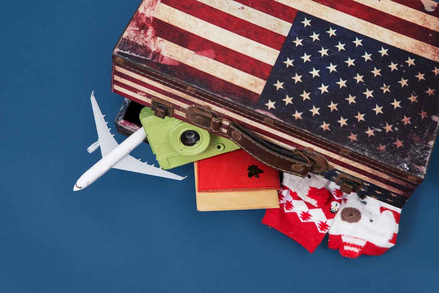 A suitcase featuring the American flag, with items like a toy airplane, a green camera, a red passport, and holiday socks sticking out, symbolizing travel, immigration, and holiday spirit.