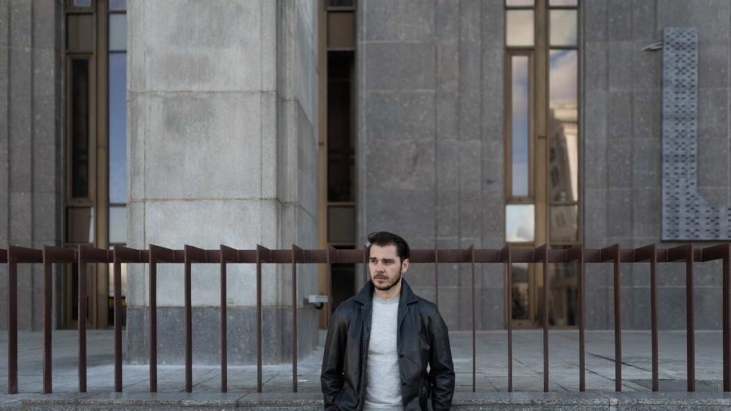 Man in a brown coat holding a microphone in front of a historical building with columns, symbolizing authority and public speaking.