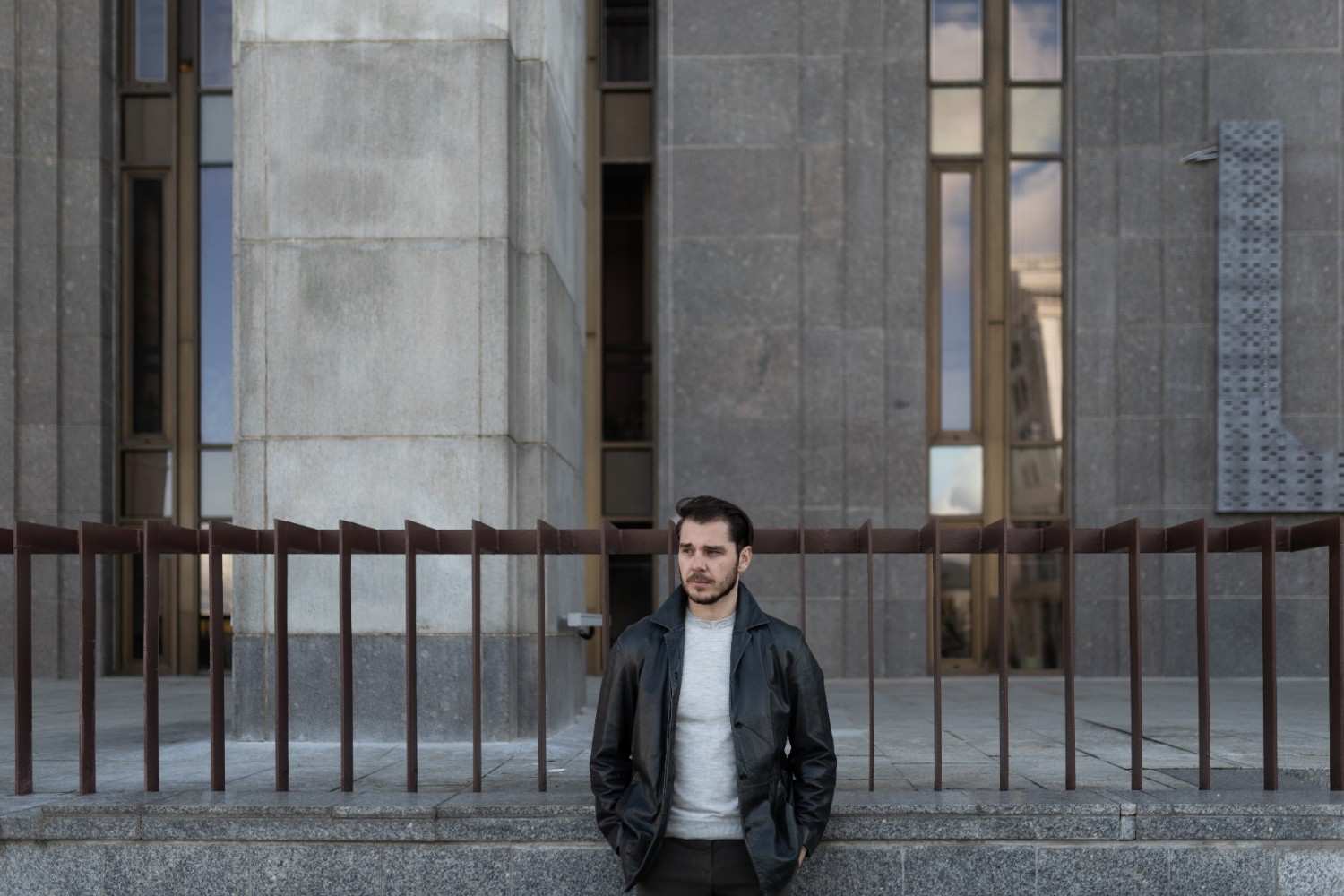 Man in a brown coat holding a microphone in front of a historical building with columns, symbolizing authority and public speaking.