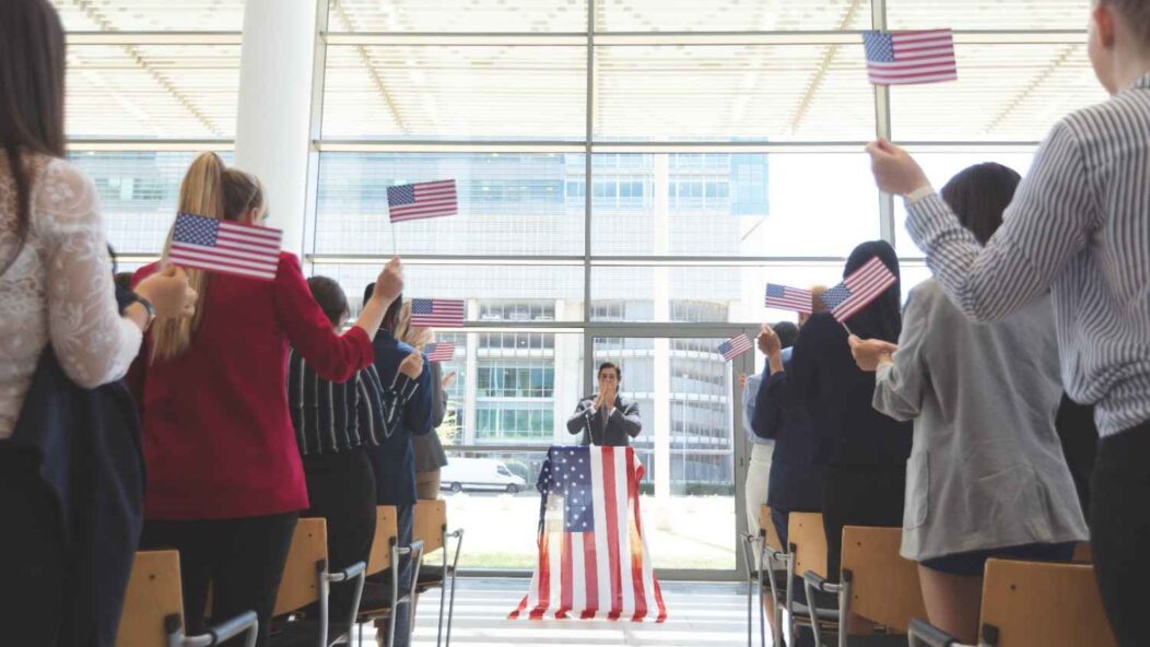 New citizens at a naturalization ceremony in the United States, raising their hands to take the Oath of Allegiance with U.S. flags in the foreground.