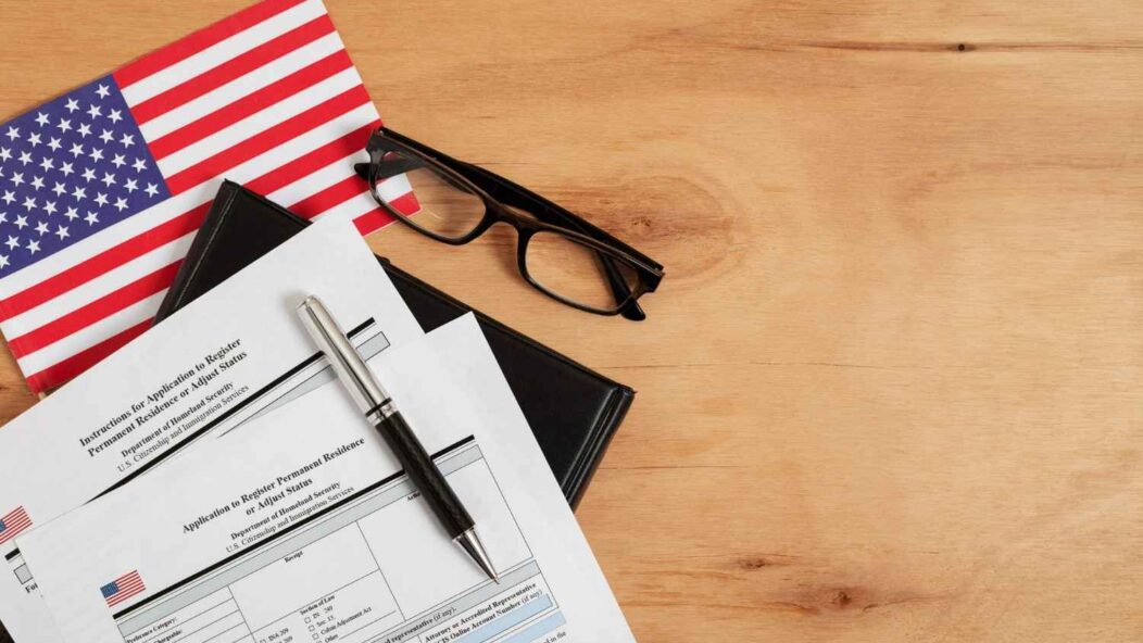 USCIS Form I-485 application papers, pen, glasses, and an American flag on a wooden table, representing the updated guidelines.