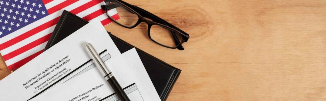 USCIS Form I-485 application papers, pen, glasses, and an American flag on a wooden table, representing the updated guidelines.