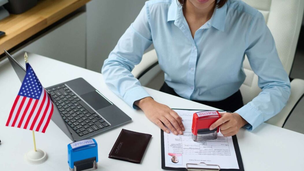 Young woman surprised with a phone in hand, standing in front of the U.S. flag, symbolizing visa-related queries and challenges.