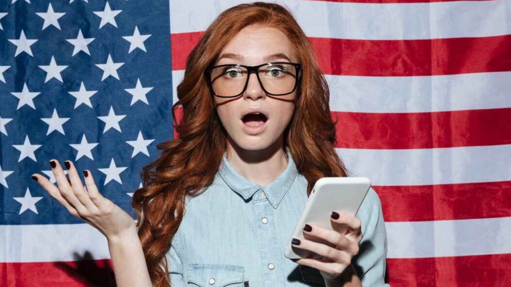 Young woman surprised with a phone in hand, standing in front of the U.S. flag, symbolizing visa-related queries and challenges.