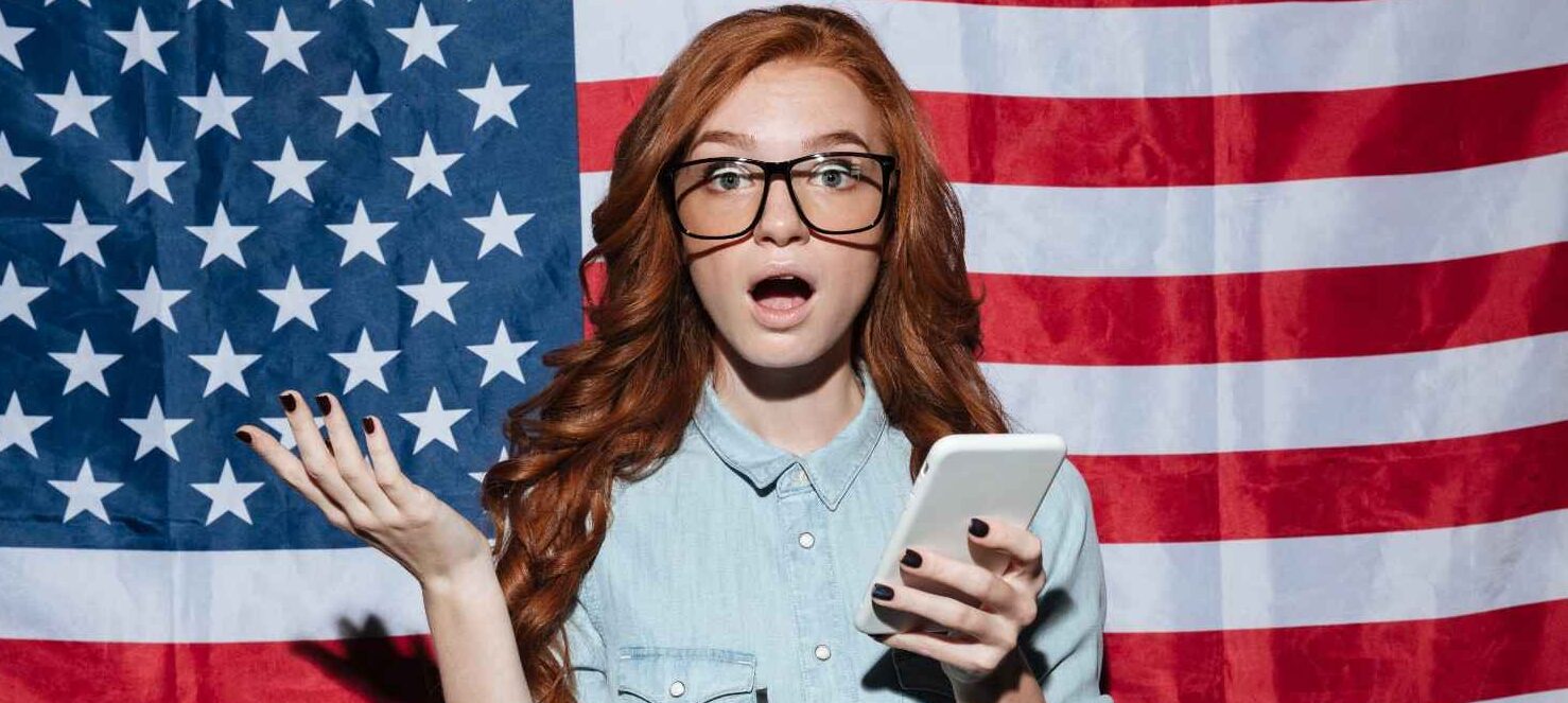 Young woman surprised with a phone in hand, standing in front of the U.S. flag, symbolizing visa-related queries and challenges.