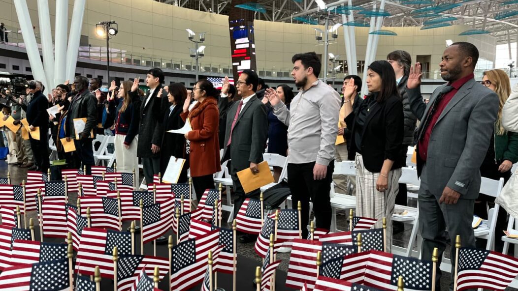 New citizens at a naturalization ceremony in the United States, raising their hands to take the Oath of Allegiance with U.S. flags in the foreground.