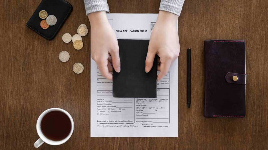 Hands holding a passport over a visa application form with coins, a wallet, pen, and coffee cup on a wooden table.