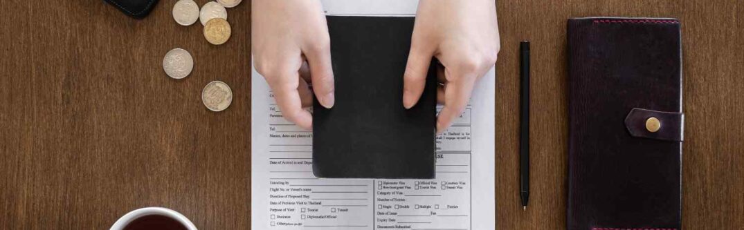 Hands holding a passport over a visa application form with coins, a wallet, pen, and coffee cup on a wooden table.