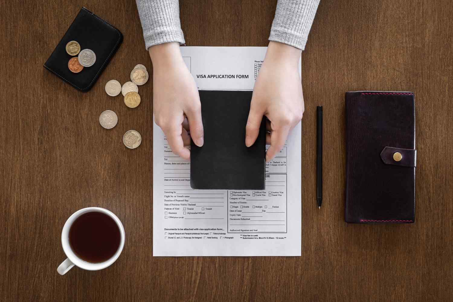 Hands holding a passport over a visa application form with coins, a wallet, pen, and coffee cup on a wooden table.