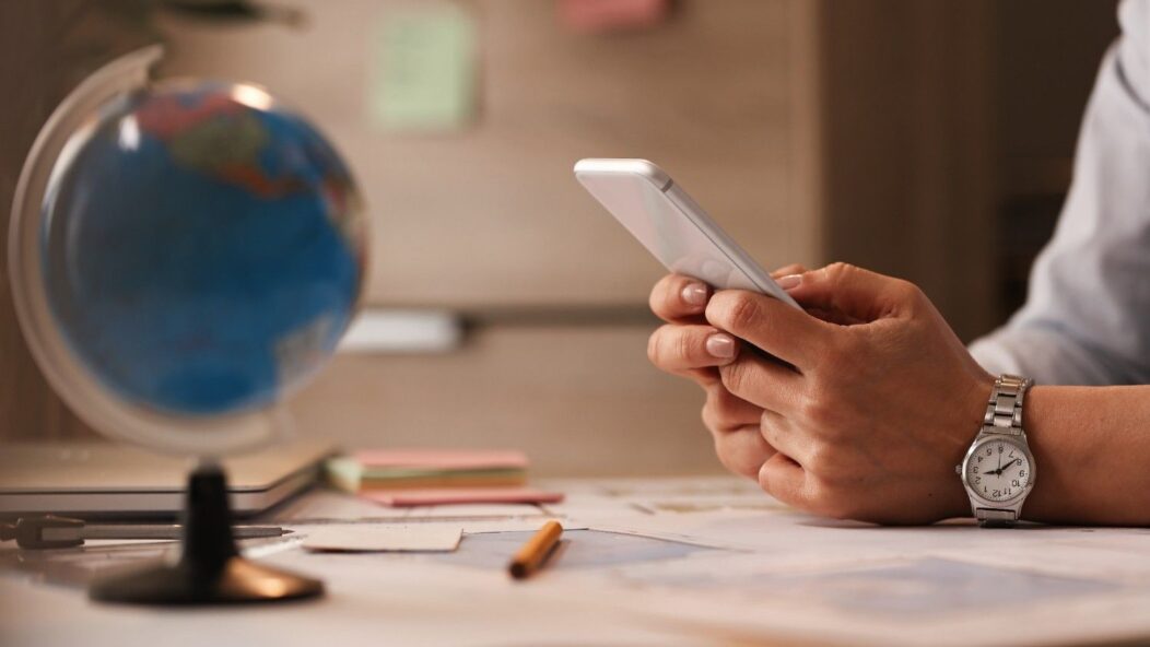 Person reviewing social media on smartphone, with a globe and documents in the background, symbolizing USCIS social media policy for immigration applications.