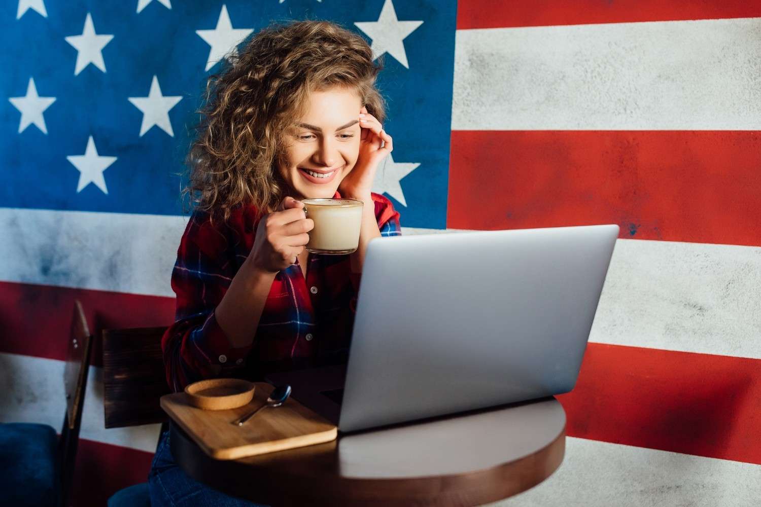 Smiling woman with curly hair enjoying coffee while working on a laptop with the American flag in the background, reflecting H-2A Program updates.